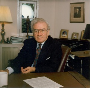An older man in a black suit sits behind a desk in an office