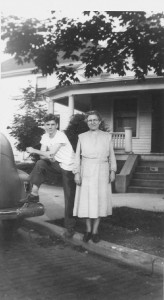 A young boy and an older woman standing outside a house under a tree next to a parked car. The boy has his foot up on the car's rear bumper.