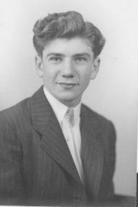 A young man poses for a headshot-style photograph, wearing a white shirt and a pinstripe black suit