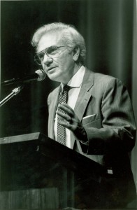 A man in a gray suit and tie stands behind a podium and speaks into a microphone. He gestures with his left hand.